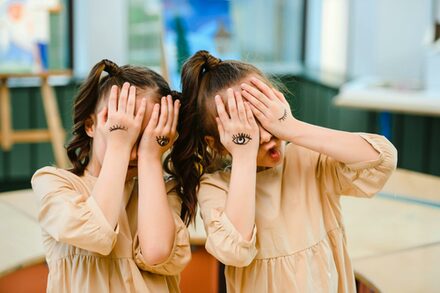 zwei Mädchen halten ihre Hände vor das Gesicht, auf dem Handrücken sind Augen gemalt