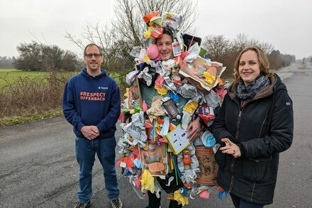 Ein mit Müll behängter Mann, der "Mülli" verkörpert, steht zwischen zwei Personen