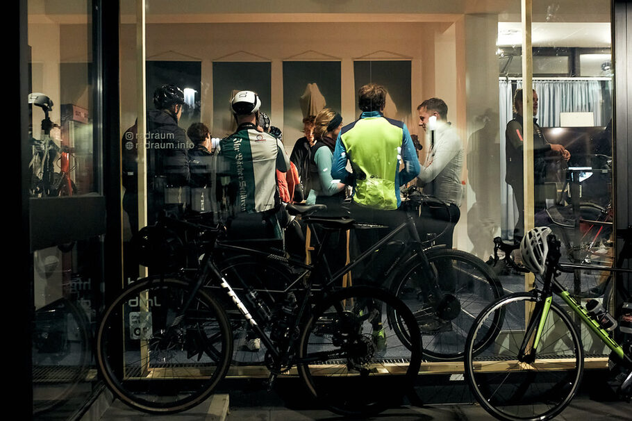 Durch eine Fensterscheibe sieht man Menschen in einem Raum, davor stehen Fahrräder.