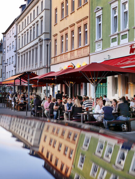 Menschen sitzen im Sommer an Tischen vor Cafes und Restaurants am Wilhelmsplatz.