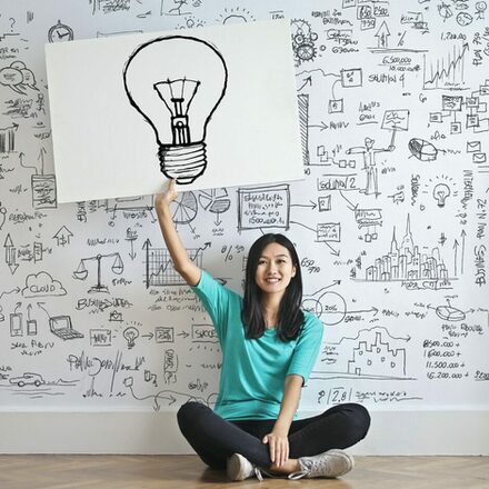 Frau sitzt vor einer Wand mit Notizen und hält ein Schild mit einer Glühbirne hoch