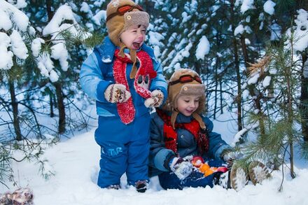 zwei Kinder in Schneeanzügen spielen unter einer Tanne im Schnee