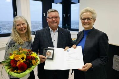 Rainer Marx hält die Bürger-Medaille in der Hand, links steht seine Ehefrau Gerda Marx mit einem Blumenstrauß, rechts von ihm Bürgermeisterin Sabine Groß.