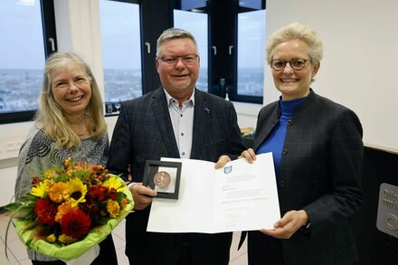 Rainer Marx hält die Bürger-Medaille in der Hand, links steht seine Ehefrau Gerda Marx mit einem Blumenstrauß, rechts von ihm Bürgermeisterin Sabine Groß.