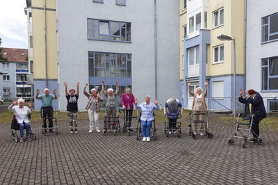 Seniorinnen und Senioren stehen mit ihren Rollatoren auf einem Platz und heben die Arme.