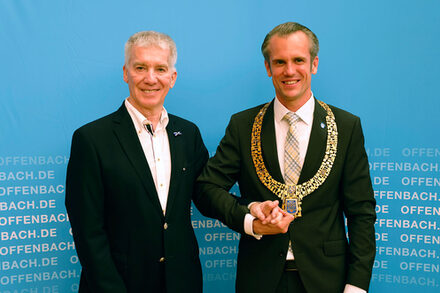 Alfred Clouth (links) steht Hand in Hand mit Oberbürgermeister Felix Schwenke nach der Vergabe der Ehrenplakette der Stadt vor einer Leinwand.