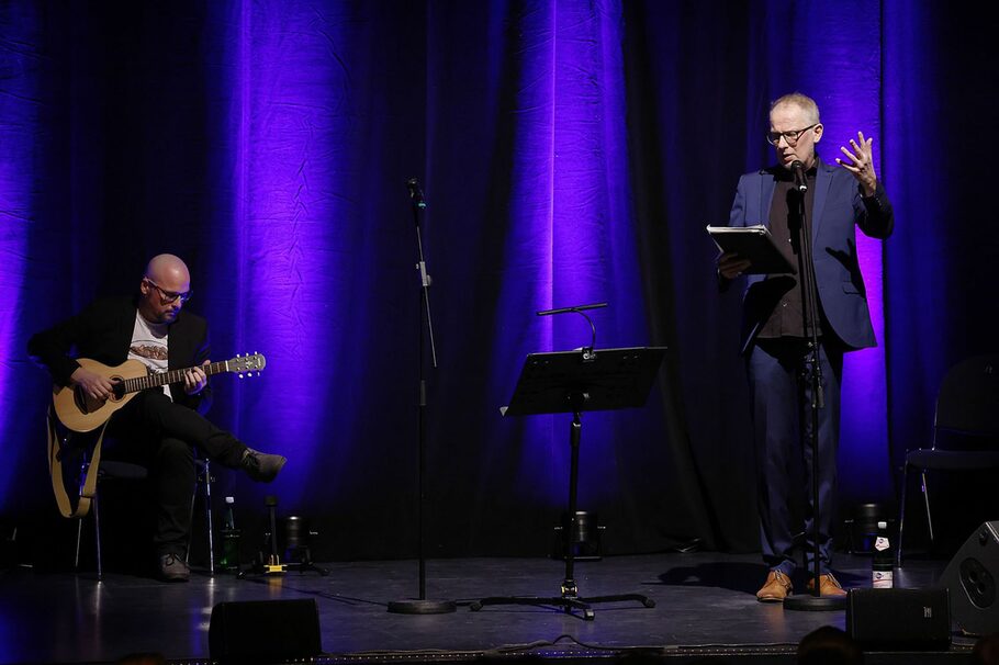 Zwei Männer auf der Bühne, einer sitzt und spielt Gitarre, der andere steht am Mikrofon und spricht.