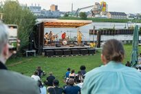 Eine Band spielt auf einer Bühne, die am Mainufer aufgebaut ist. Menschen sitzen im Gras und hören zu.