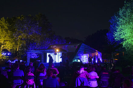 Besucher sitzen während einer Veranstaltung vor dem blau erleuchtetem Besucherzentrum im Wetterpark.
