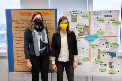 Zwei Frauen mit Maske stehen vor zwei Pinnwänden mit Texten und Bildern.