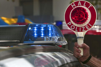 Eine Hand hält eine Kelle mit der Aufschrift "Halt Polizei" neben einem Blaulicht eines Polizeiautos.