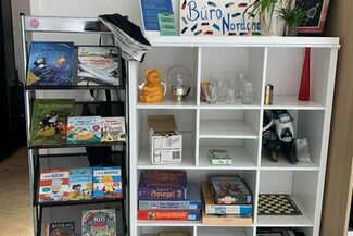 Ein Regal mit verschiedenen Gegenständen wie Brettspiele und Deko-Artikeln, daneben steht ein Regal mit Kinderbüchern.
