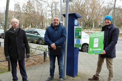 Drei Herren am Parkscheinautomat, einer hält ein Schild zum Handyparken