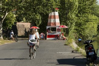 Blick auf einen kleinen Kiosk in Form eines Leuchtturmes, mit Radfahrern und Spaziergängern.