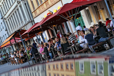 Menschen sitzen im Sommer an Tischen vor Cafes und Restaurants am Wilhelmsplatz.