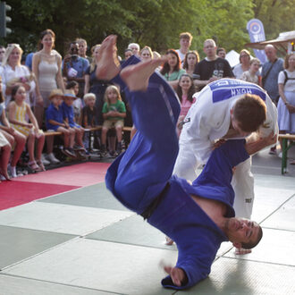 Zwei Personen beim Judokampf, drumherum Publikum.
