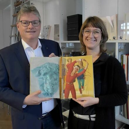 Das Bild zeigt den Stadtwerke-Geschäftsführer Peter Walther und die Leiterin des Klingspor Museums, Dr. Dorothee Ader, bei der Übergabe des Künstlerbuchs "Enorm in Form". Auf der aufgeschlagenen Seite des Buchs sind Frauen im Aerobic-Dress zu sehen.
