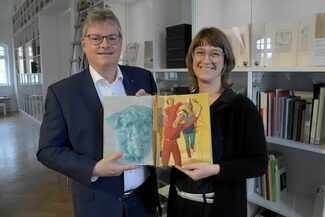 Das Bild zeigt den Stadtwerke-Geschäftsführer Peter Walther und die Leiterin des Klingspor Museums, Dr. Dorothee Ader, bei der Übergabe des Künstlerbuchs "Enorm in Form". Auf der aufgeschlagenen Seite des Buchs sind Frauen im Aerobic-Dress zu sehen.
