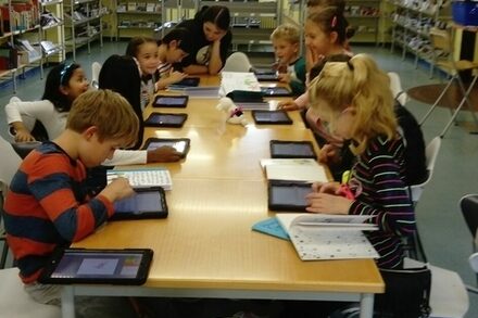 Kinder sitzen an einem Tisch und lernen mit Tablets und Büchern.