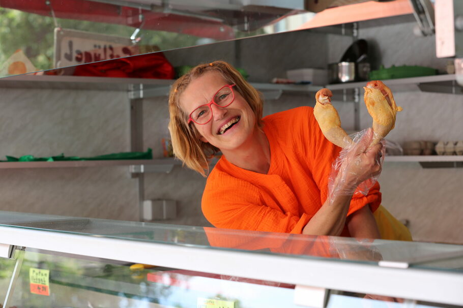 Eine lachende Frau mit orangener Bluse hält zwei Hähnchenschenkel in der Hand und steht hinter ihrer Theke.