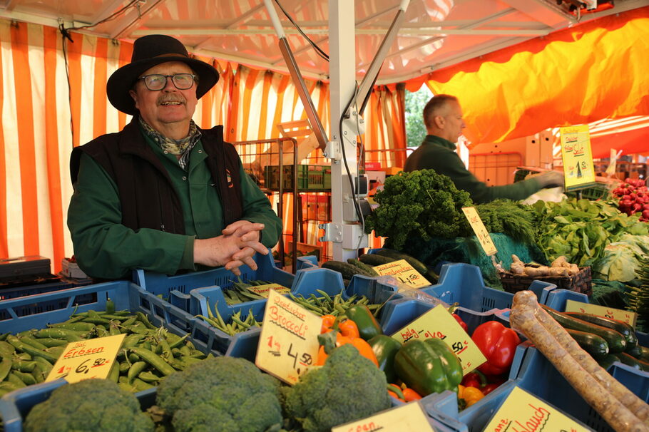 Günter Buxmann steht mit einem Filzhut hinter blauen Plastikkisten voller Gemüse.