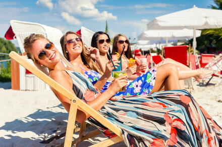 Vier lachende Frauen mit Cocktails in Liegestühlen an einem Strand.