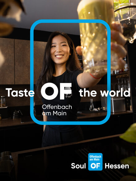 Eine Frau in schwarzem T-Shirt steht hinter einer Küchentheke und hält ein Glas in die Kamera. Auf dem Bild liegt das Offenbach Logo sowie der Kampagnen-Slogan.
