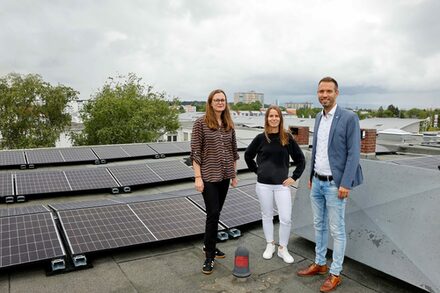 Zwei Frauen und ein Mann stehen auf einem Hausdach vor einer Photovoltaikanlage.