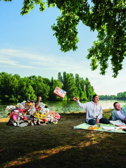 Drei Personen sitzen auf einer Pickenick-decke am Mainufer, eine wirft Müll auf einen als Müllberg verkleideten Schauspieler