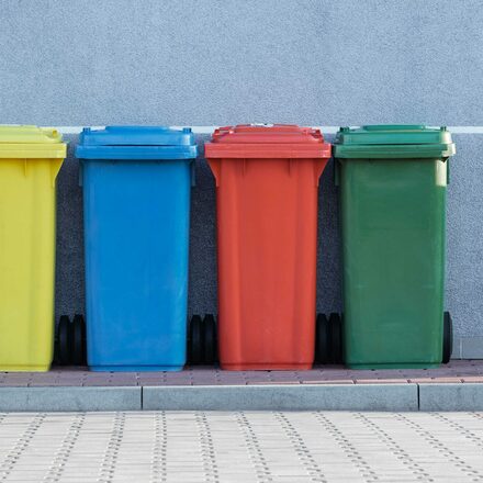 Eine gelbe, eine  blaue, eine rote und eine grüne Mülltonne stehen in einer Reihe vor einer Hauswand.