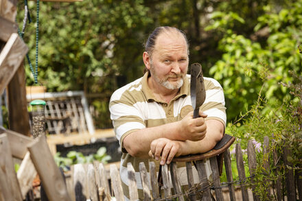 Ein Mann lehnt an einem Gartenzaun und hat eine Schaufel in der Hand