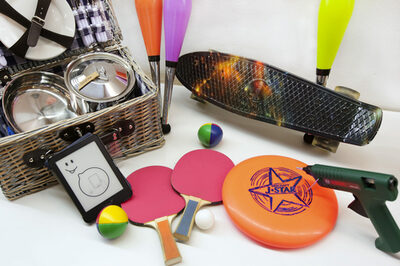 Auf einem Tisch liegen: Ein Skateboard, ein Picknick-Korb, Tischtennisschläger, eine Heißklebepistole, ein E-Reader, ein Frisbee und Jonglierstäbe.