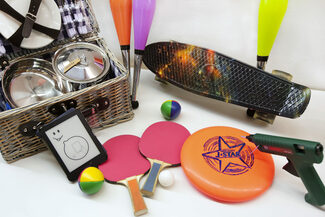 Auf einem Tisch liegen: Ein Skateboard, ein Picknick-Korb, Tischtennisschläger, eine Heißklebepistole, ein E-Reader, ein Frisbee und Jonglierstäbe.