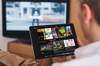 Ein Mann betrachtet auf einem Tablett eine Auswahl vorgeschlagener Filmtitel mit Fotos an an, im Hintergrund ein Fernseher.