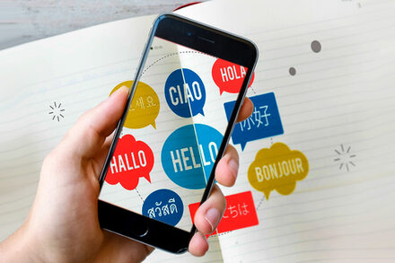 Eine Hand hält eine Handy über einem Heft, in dem das Wort Hallo in verschiedenen Sprachen steht.