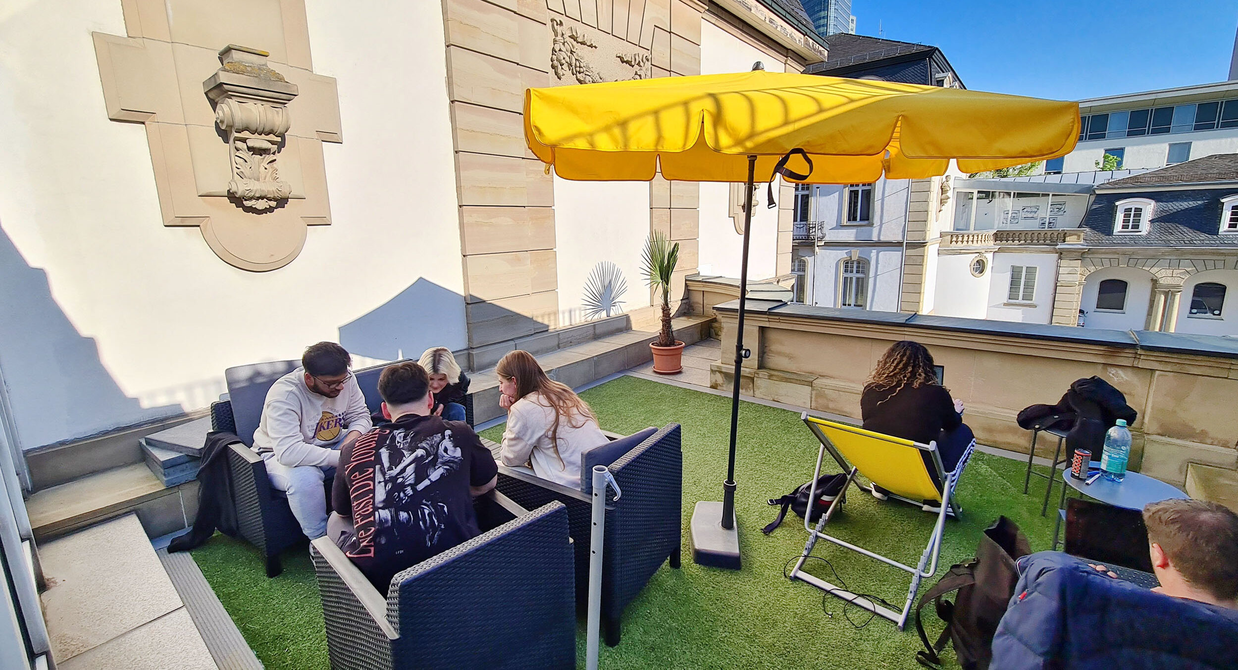 Jugendliche sitzen auf einer kleinen Terrasse, in der Mitte steht ein gelber Sonnenschirm.