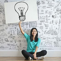 Junge Frau sitzend hält weißes Plakat mit einer Glühbirnenzeichnung darauf in der Hand