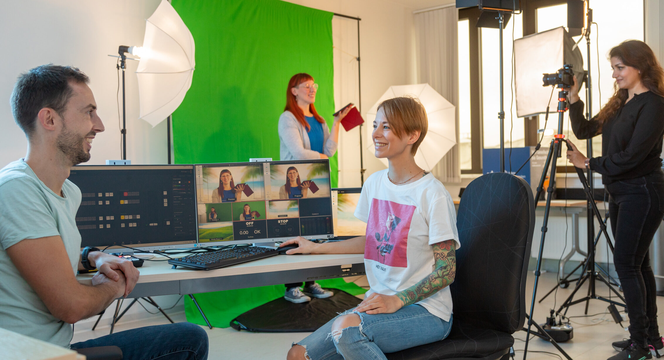 An einem Tisch sitzen zwei Personen vor einem Computer. Im Hintergrund steht eine Frau mit Kamera, die eine andere Frau filmt.