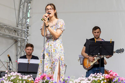Eine Frau singt in ein Mikrofon, im Hintergrund sind zwei weitere Musiker zu sehen.