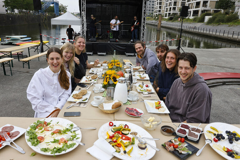 Sieben junge Menschen sitzen beim Frühstück an der festlich gedeckten Tafel auf der Hafentreppe.