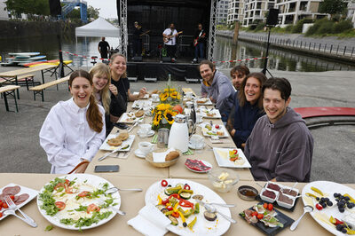 Sieben junge Menschen sitzen beim Frühstück an der festlich gedeckten Tafel auf der Hafentreppe.