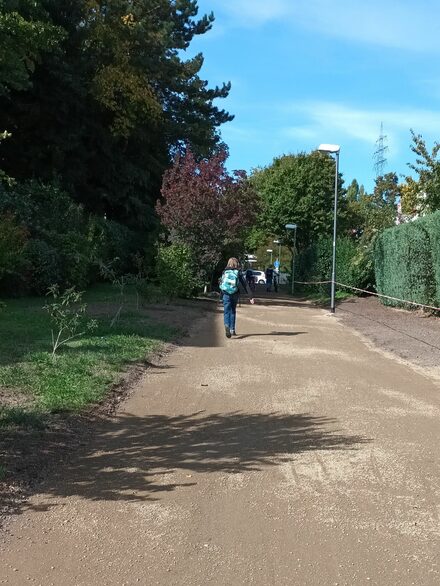 Ein breiter brauner Kiesweg, ein Junge läuft darauf, im Hintergrund grüne Laubbäume.