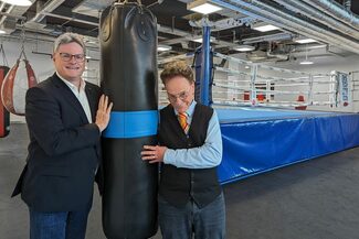 Zwei Männer stehen in einer Boxhalle, zwischen ihnen hängt ein Boxsack.